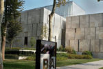 Modern architectural building with stone exterior, surrounded by trees and landscaped greenery. A sign with the letters "BA" is in the foreground.