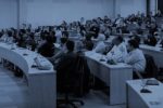 A lecture hall with a speaker addressing a seated audience. The room is arranged with curved seating and attendees appear engaged, some taking notes.