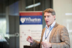 A person speaking in front of a Wharton conference banner, wearing a checkered shirt and brown blazer, holding a conversation or presentation.