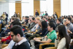 A large crowd of people sitting in chairs, attentively listening, possibly at a seminar or conference. The setting appears to be an indoor venue with natural light.