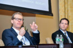 Two people seated on a panel at a conference, with one speaking and gesturing with their hand while holding a microphone. Water bottles are on the table in front of them.