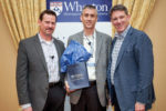 Three individuals in business attire stand in front of a Wharton School banner. One holds a gift bag from the Wharton Store. They are smiling.