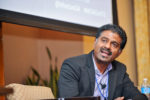 A person in a suit speaks while seated at a conference or panel discussion, wearing a microphone headset.