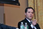 A person in a suit is speaking and gesturing with their hand, seated at a conference panel. Two water bottles are in the foreground.