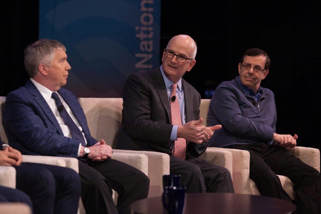 Three people are seated on a panel during a discussion or conference. The individual in the center appears to be speaking while gesturing with their hands.