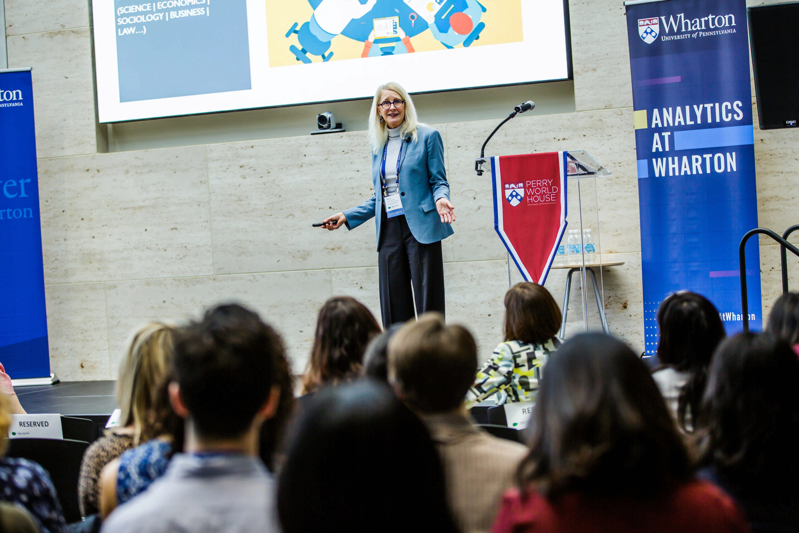 A person in professional attire is giving a presentation at an event, possibly related to analytics at Wharton, with an audience listening attentively.