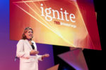 A person wearing a white blazer is speaking on stage during a TED@WellsFargo event, with a large "ignite" sign in the background.