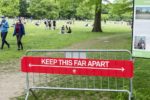 A park setting with people scattered on the grass, maintaining distance. A red sign on a barrier reads "Keep This Far Apart," indicating social distancing measures.