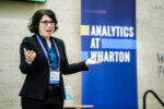 A person speaking at a conference, wearing a speaker badge and a lanyard. The background features a banner that reads "Analytics at Wharton."