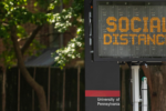 An electronic sign displaying the message "SOCIAL DISTANCE" is mounted above a pole with "University of Pennsylvania" written on it, surrounded by trees.