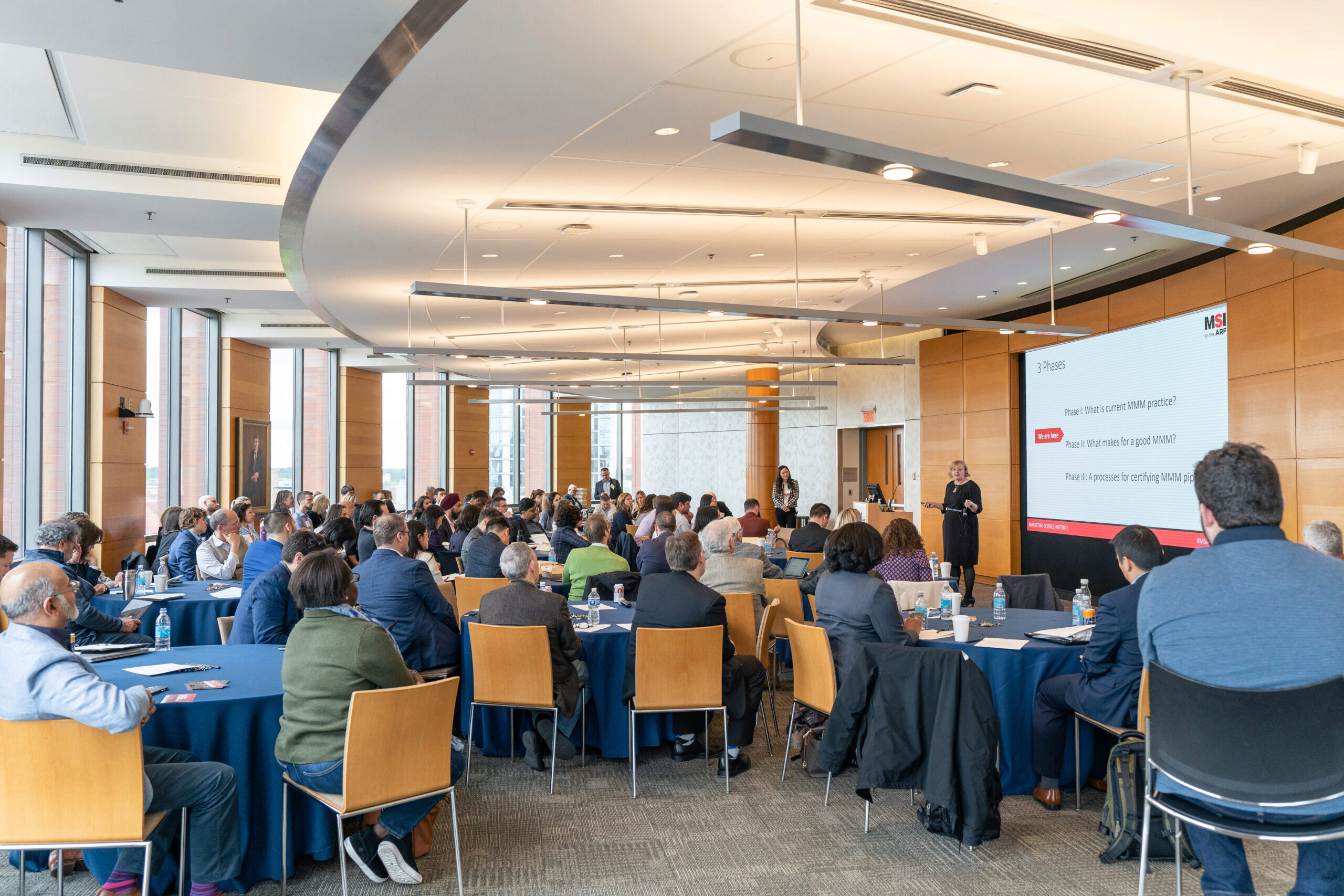 A conference room filled with people seated at round tables, listening to a speaker at the front next to a large presentation screen.