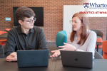 Two individuals in a modern office setting, engaged in conversation with laptops open in front of them. The backdrop features a brick wall and the text "Analytics at Wharton."