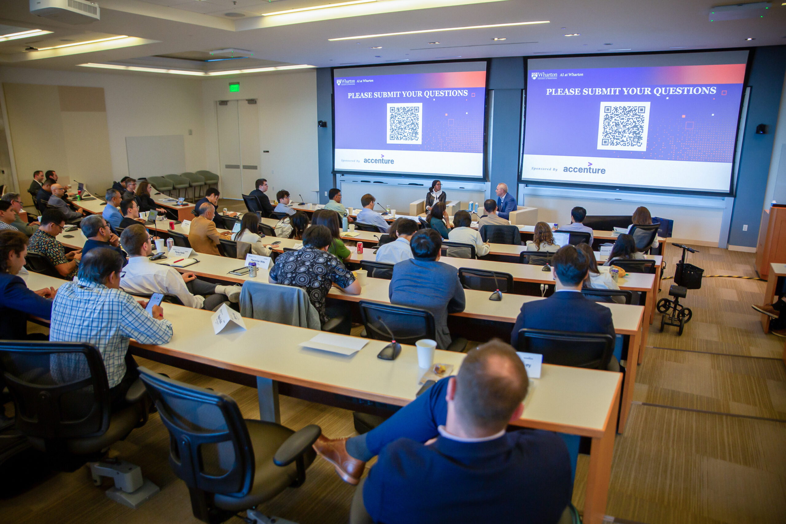 A group of people are seated in a lecture hall, attending a presentation. Two large screens display the text "PLEASE SUBMIT YOUR QUESTIONS" with a QR code. The setting suggests a professional or academic seminar.