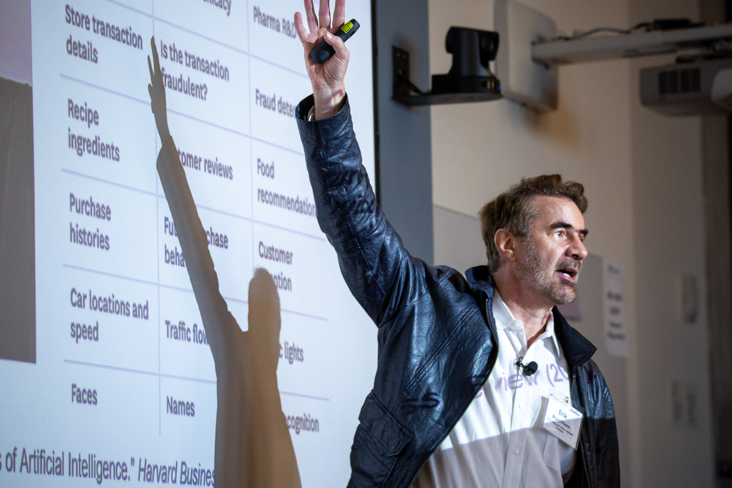 A person wearing a leather jacket is giving a presentation, gesturing towards a projected slide with a table of text about artificial intelligence.