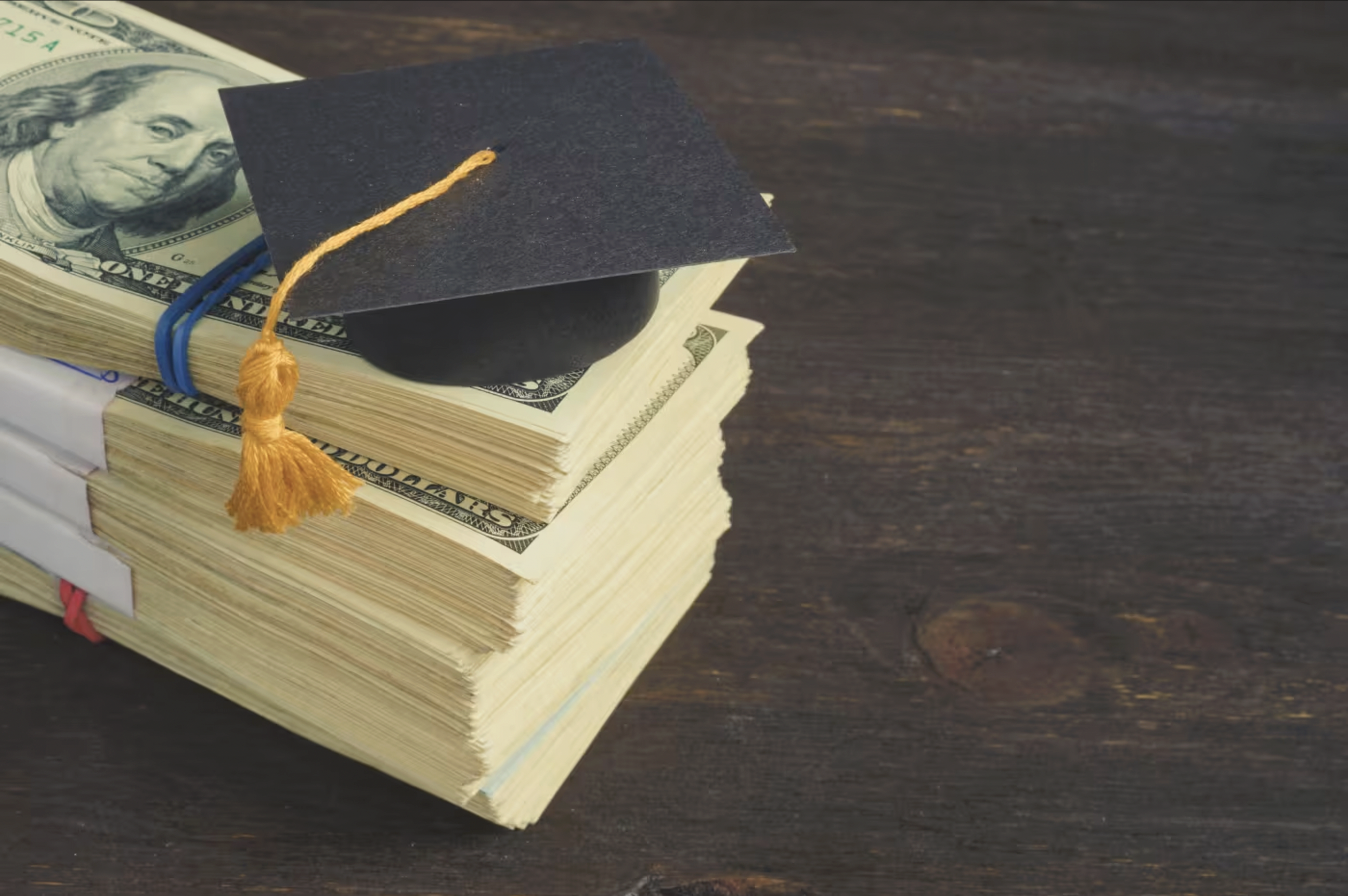 Stack of U.S. hundred-dollar bills with a black graduation cap on top, symbolizing student loans or the cost of education.