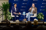 Two people sit on stage speaking at a panel discussion, in front of a Wharton School backdrop with plants on either side.