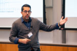 A person wearing glasses and a name badge is speaking during a presentation, gesturing with one hand. A projected screen with a graph is visible in the background.