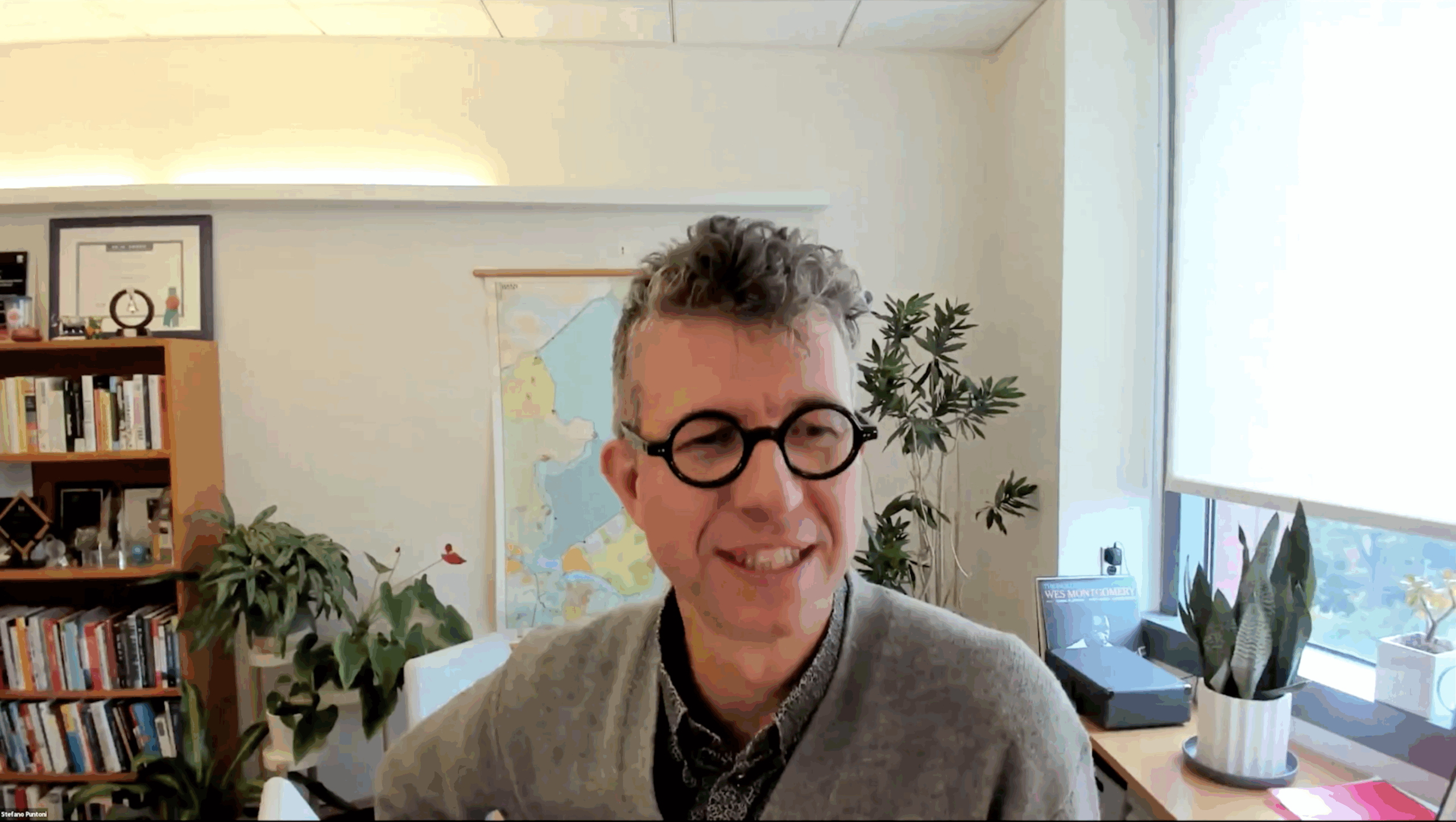 A person with glasses sitting in a home office, surrounded by plants, books, and a large wall map in the background.