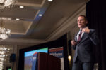 A person in a suit giving a presentation in a conference room with chandeliers. There are Wharton banners and a podium in the background.