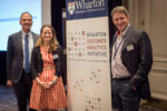 Three individuals pose near a banner for the Wharton Customer Analytics Initiative from the University of Pennsylvania, highlighting business transformation through customer data.