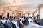 A conference room with people seated at round tables listening to a person presenting on a stage. The room features chandeliers and elegant decor.