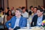 People seated at a conference, attentively listening, with notepads and water bottles on the table; a professional event setting.