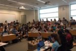 A full classroom with students seated in tiers, many using laptops, suggesting a lecture or academic setting.