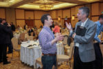 Two people are engaged in conversation at a conference or seminar inside a large hall. They are surrounded by tables and other attendees.