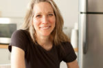 Headshot of a person with long hair, wearing a dark shirt and smiling. They are in a kitchen setting with a microwave and refrigerator in the background.