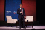 A person in a suit holds a microphone, standing between two chairs on a stage with a Wharton Customer Analytics Initiative backdrop.