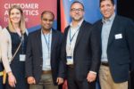 Four people are standing together, smiling, in front of banners for the Wharton University of Pennsylvania Customer Analytics Initiative. They are dressed in business attire.