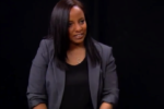 A person with long hair wearing a dark blazer and top is sitting against a black background, smiling slightly.