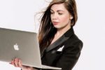 Person in a black suit holding an open laptop, looking at the screen with a focused expression. Hair is slightly windblown.