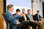 A panel discussion featuring five individuals seated in chairs, engaged in conversation. A person on the left gestures while speaking.