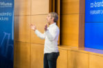 A person is giving a lecture or presentation at the Wharton School of the University of Pennsylvania. The setting includes a banner for Leadership Lectures and a screen displaying event details.