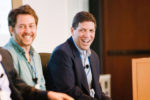 Two people are sitting and smiling during what appears to be a panel discussion or meeting. Both are dressed in business attire and wearing microphones.