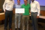 Four people stand in a classroom setting, with two of them wearing conference-style nametag shirts. They are holding a large check, indicating a presentation or award event.