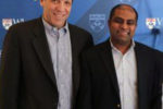 Two people in business attire stand in front of a backdrop with Wharton School logos. They are smiling, suggesting a professional event or occasion.