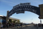 An archway sign reading "Santa Monica Yacht Harbor Sport Fishing Boating Cafes" with people walking underneath on a sunny day. The ocean and a ferris wheel are visible in the background.