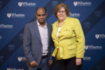 Two people standing in front of a backdrop featuring the Wharton School logo. One person is wearing a gray suit, and the other is in a yellow jacket.