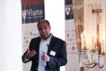 A person is giving a presentation, holding a water bottle. They are standing next to banners displaying the Wharton School logo and a decorative light fixture.